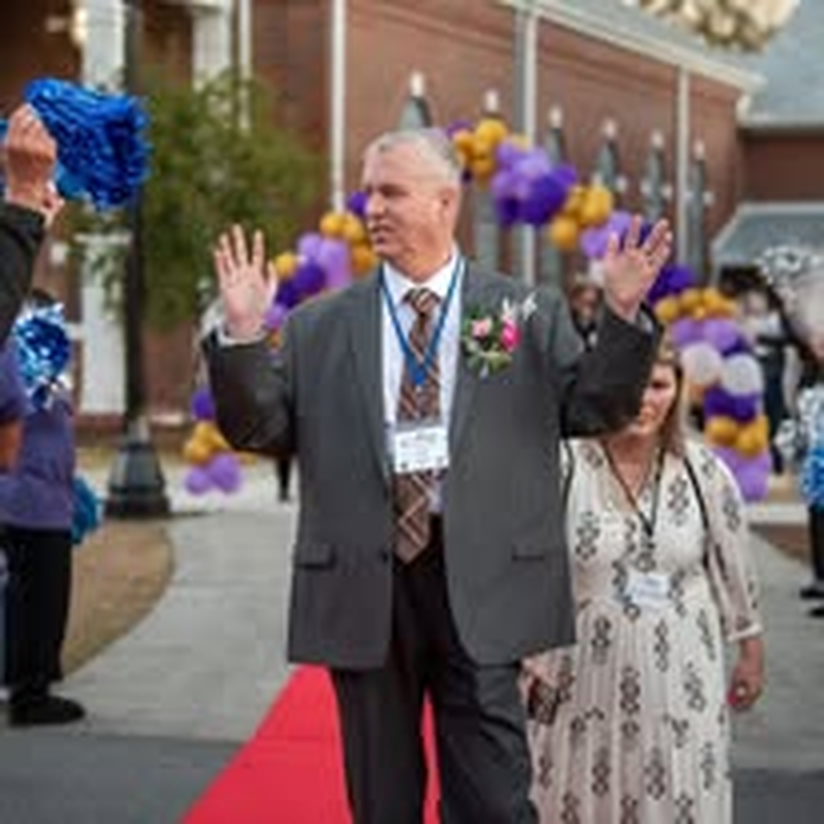 A man waving on the red carpet beneath a purple and gold balloon arch.